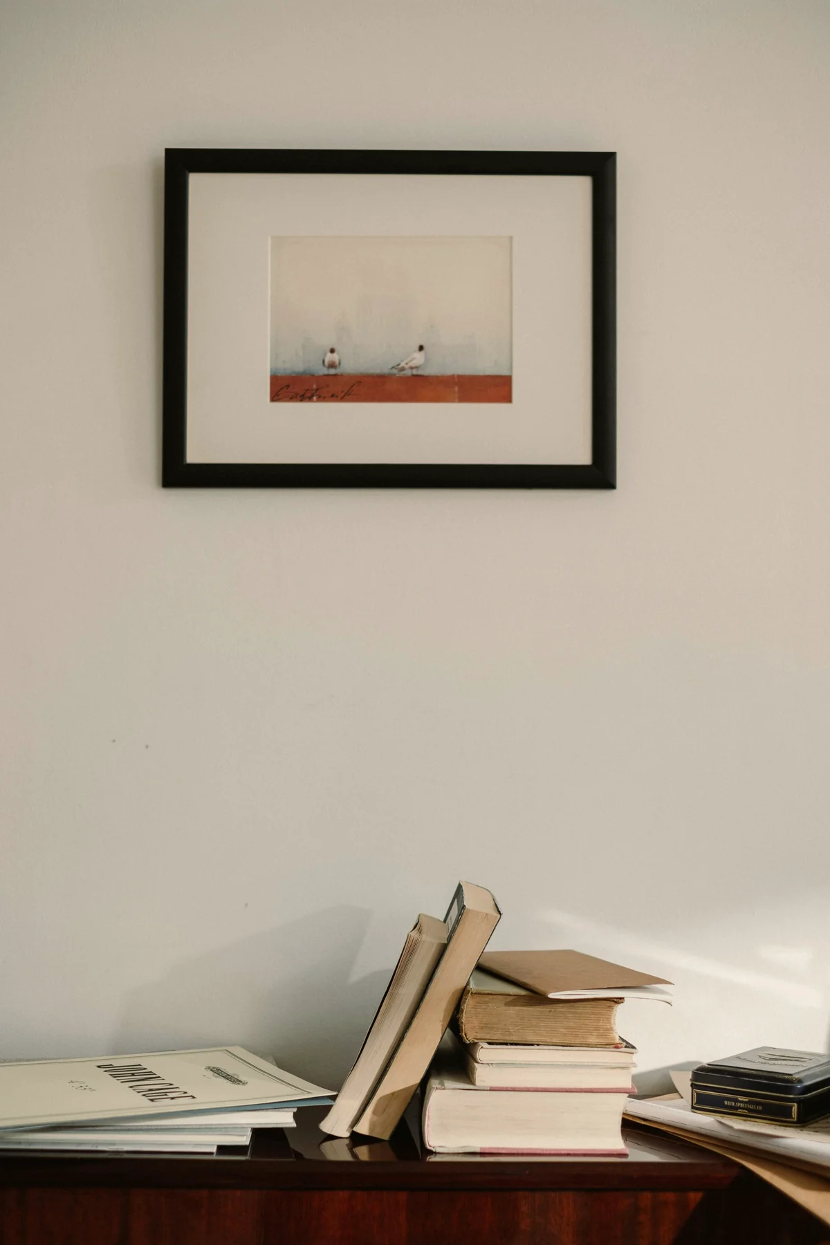 A small framed work above a wooden console — the piece is not large or small, only large or small in conversation with the books and surface beneath it.