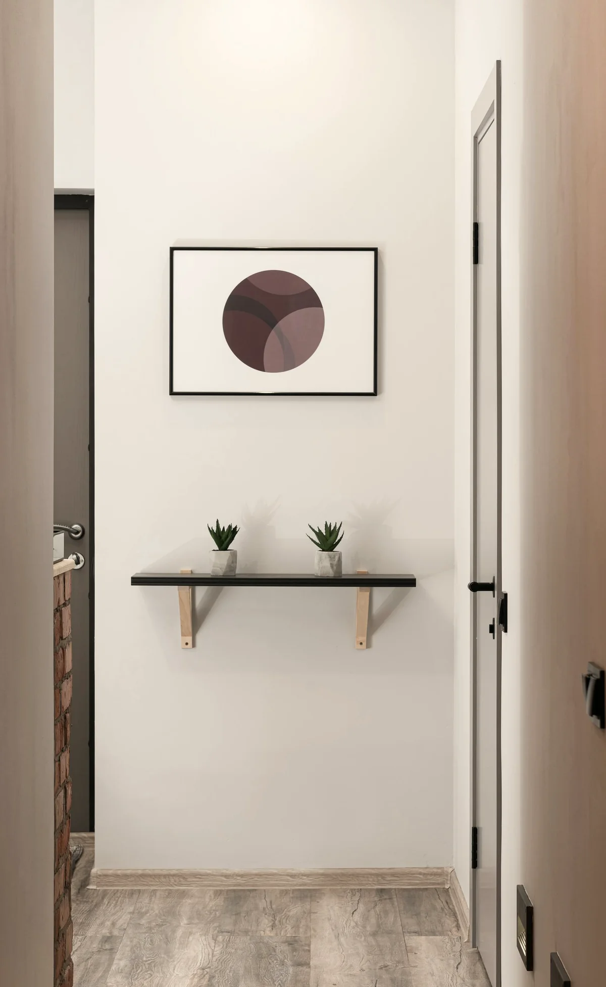 A wall that holds mostly empty space — one quiet framed work, two small plants on a slim shelf, and a long stretch of white that is part of the composition.