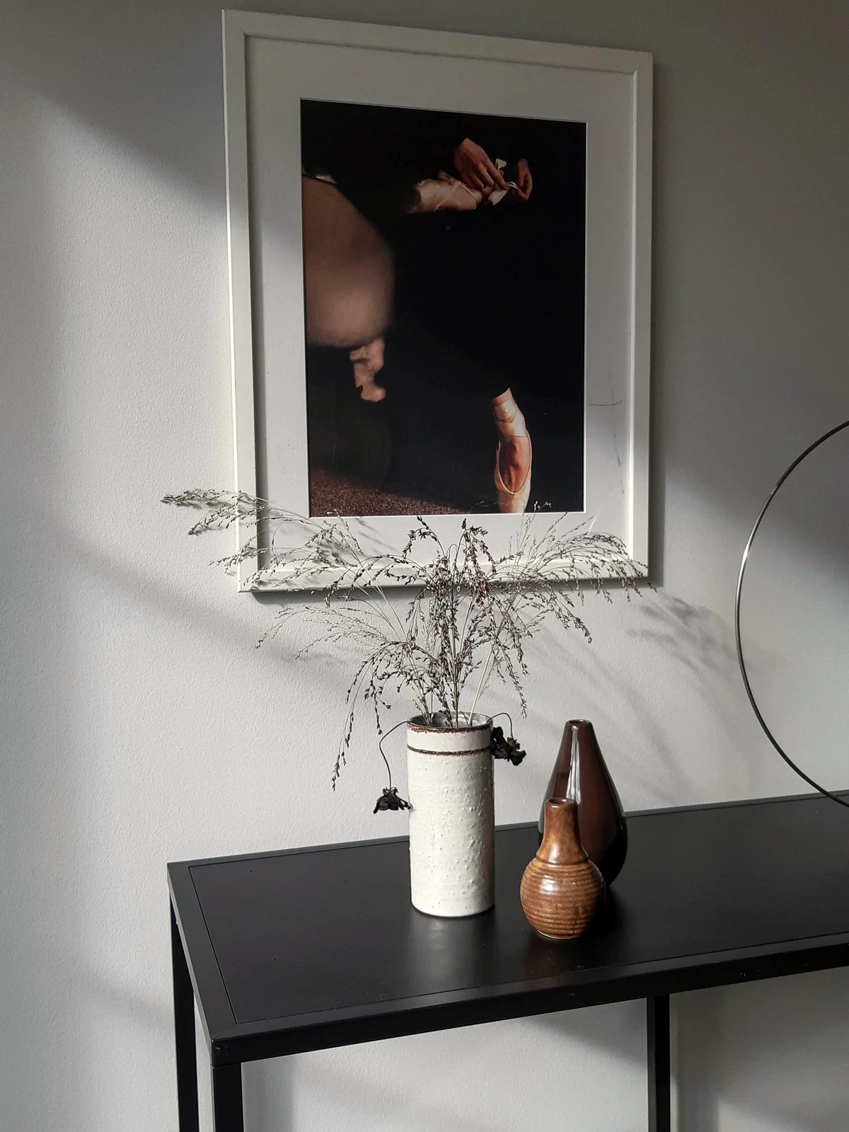 A black console holds objects from different decades — a contemporary photograph above, a hand-thrown stoneware vase, an older brown ceramic, dried grasses still arriving from the year before.